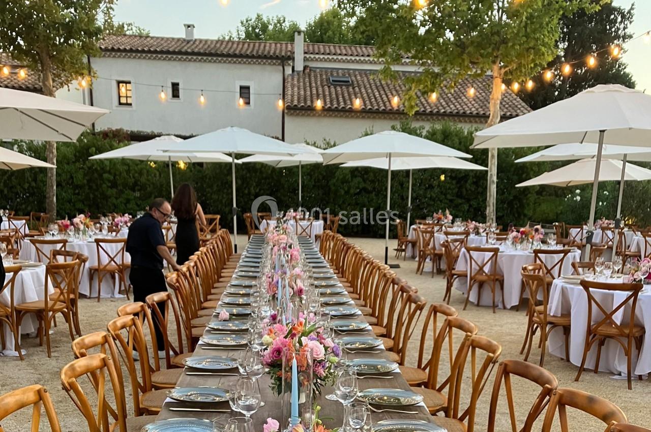 Grande table décorée de fleurs et dressée pour un repas en extérieur, entourée de petites tables sous des parasols.