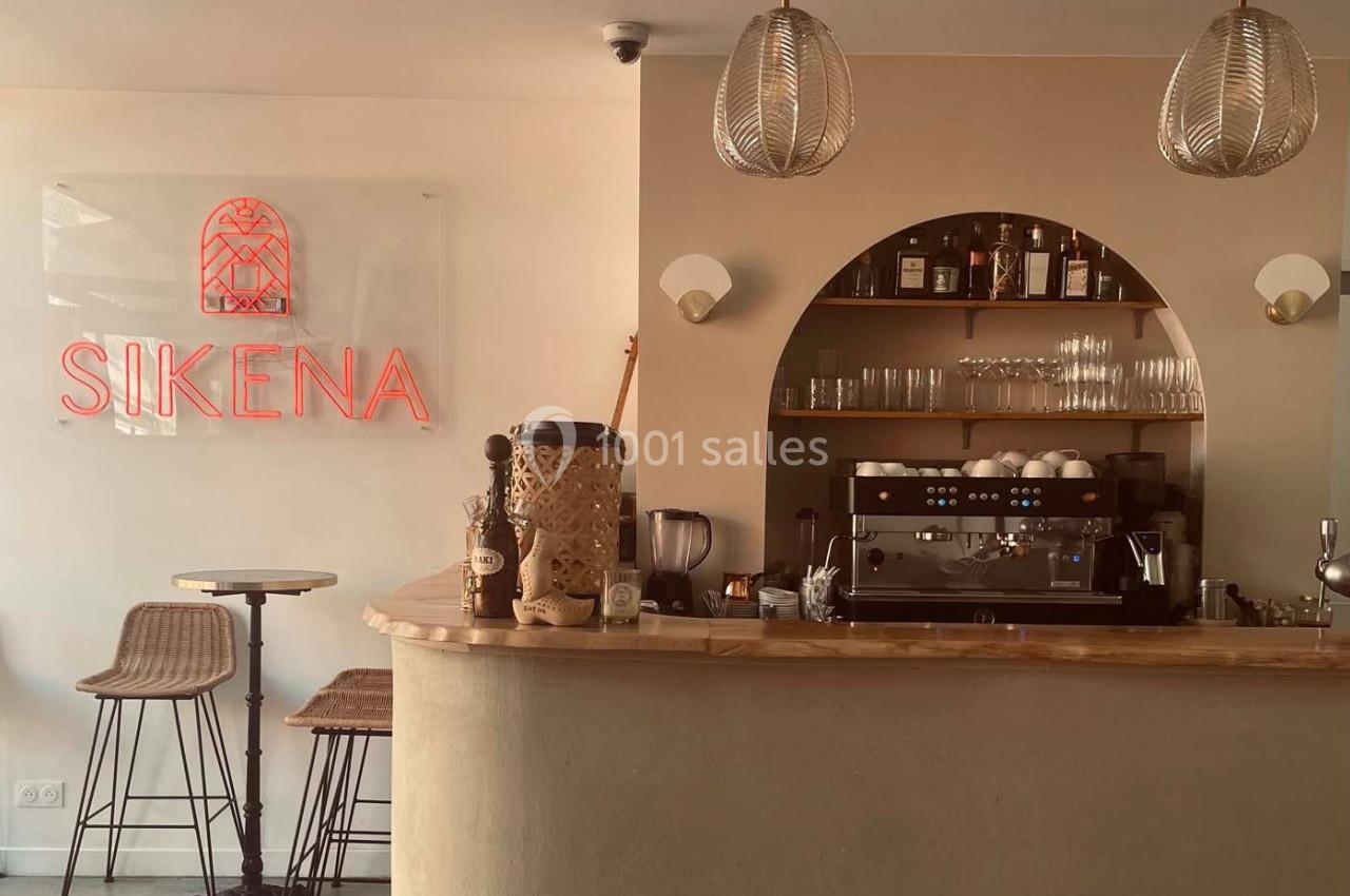 Comptoir de café avec machine à expresso, étagères garnies de bouteilles et verres, éclairage chaleureux.