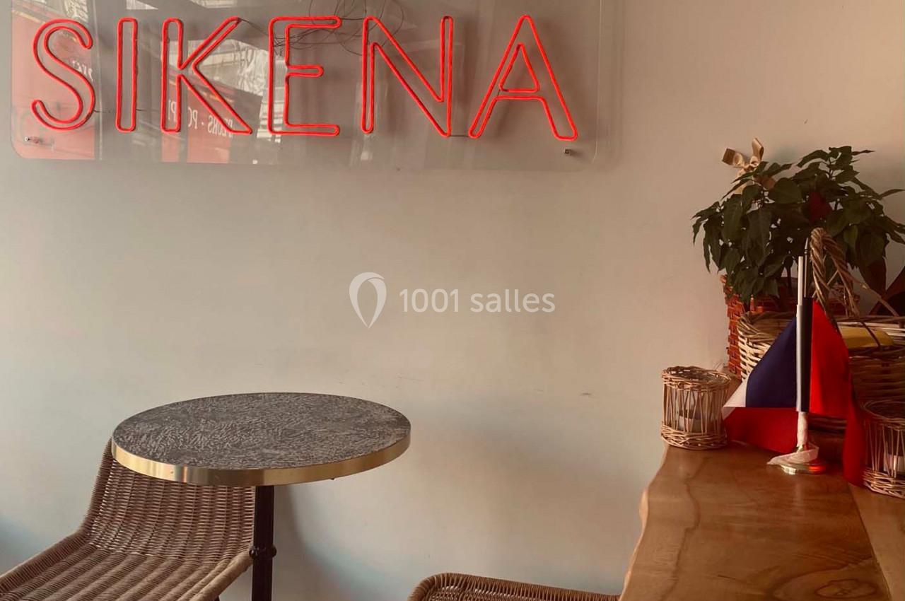 Table et chaises en rotin dans un café avec une enseigne lumineuse rouge ’SIKENA’ et un drapeau sur le comptoir.