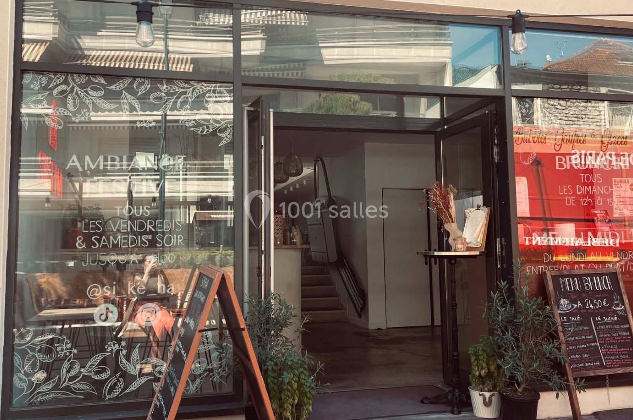 Façade vitrée d'un restaurant avec porte ouverte, ardoises d'informations et décorations végétales.