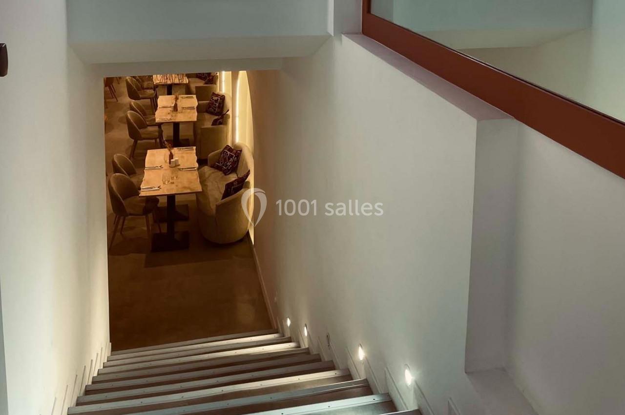 Escalier descendant vers une salle de restaurant éclairée, avec des tables dressées et des fauteuils.