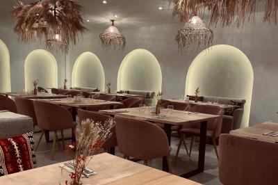 Banquette grise avec coussins rouges à motifs, tables en bois dressées, décor mural avec luminaires et ornements végétaux.