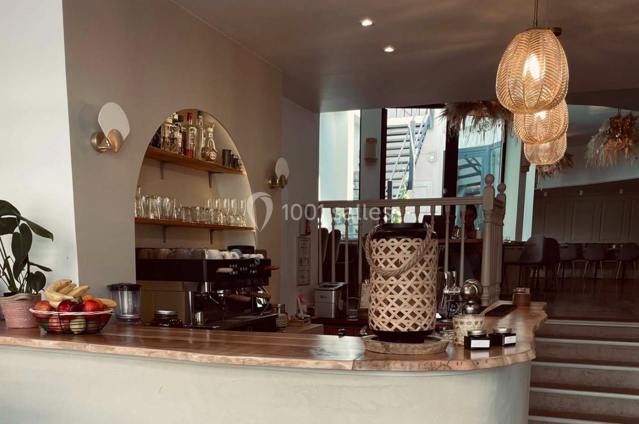 Comptoir de café avec machine à expresso, étagères de verres et bouteilles, éclairage chaleureux et décoration en rotin.