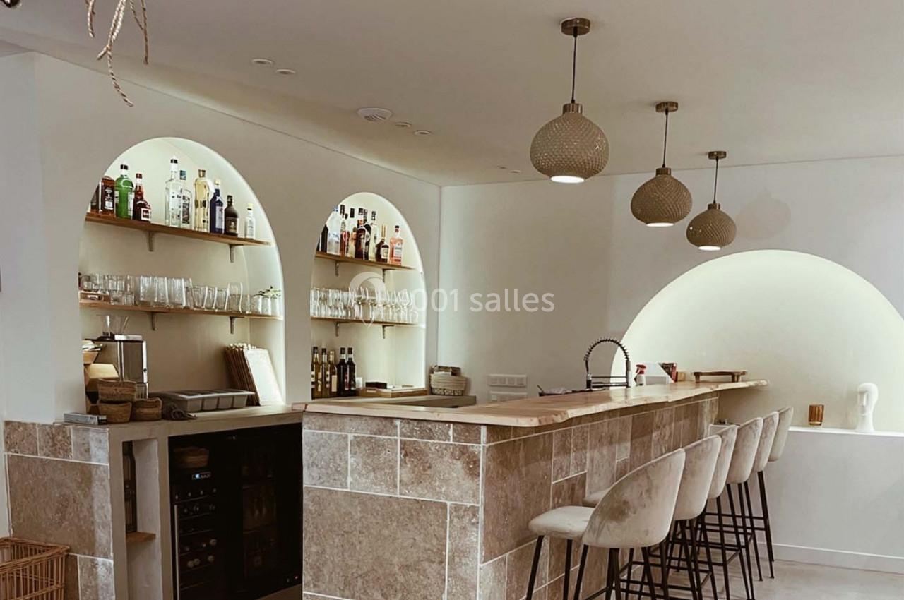 Bar moderne avec comptoir en bois, tabourets rembourrés, étagères éclairées contenant des bouteilles et des verres.