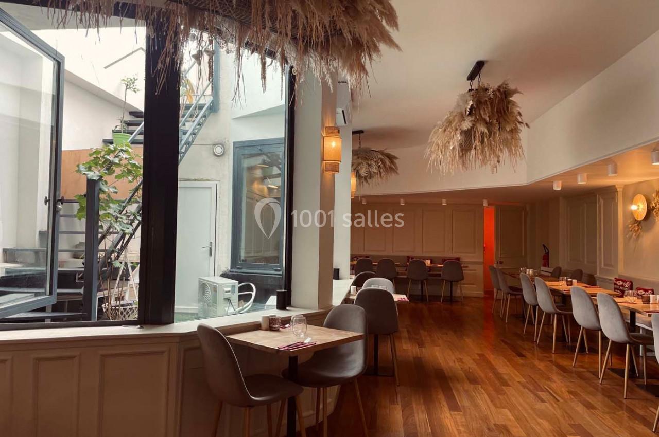Salle de restaurant lumineuse avec parquet en bois, tables dressées, chaises grises et décoration végétale suspendue.