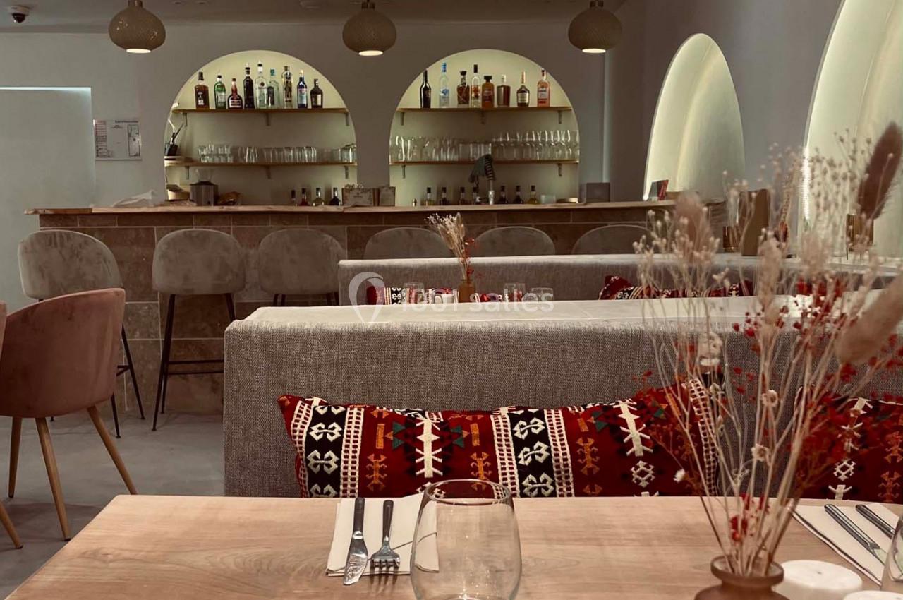 Salle de restaurant avec tables en bois, banquettes ornées de coussins à motifs et un bar en arrière-plan.