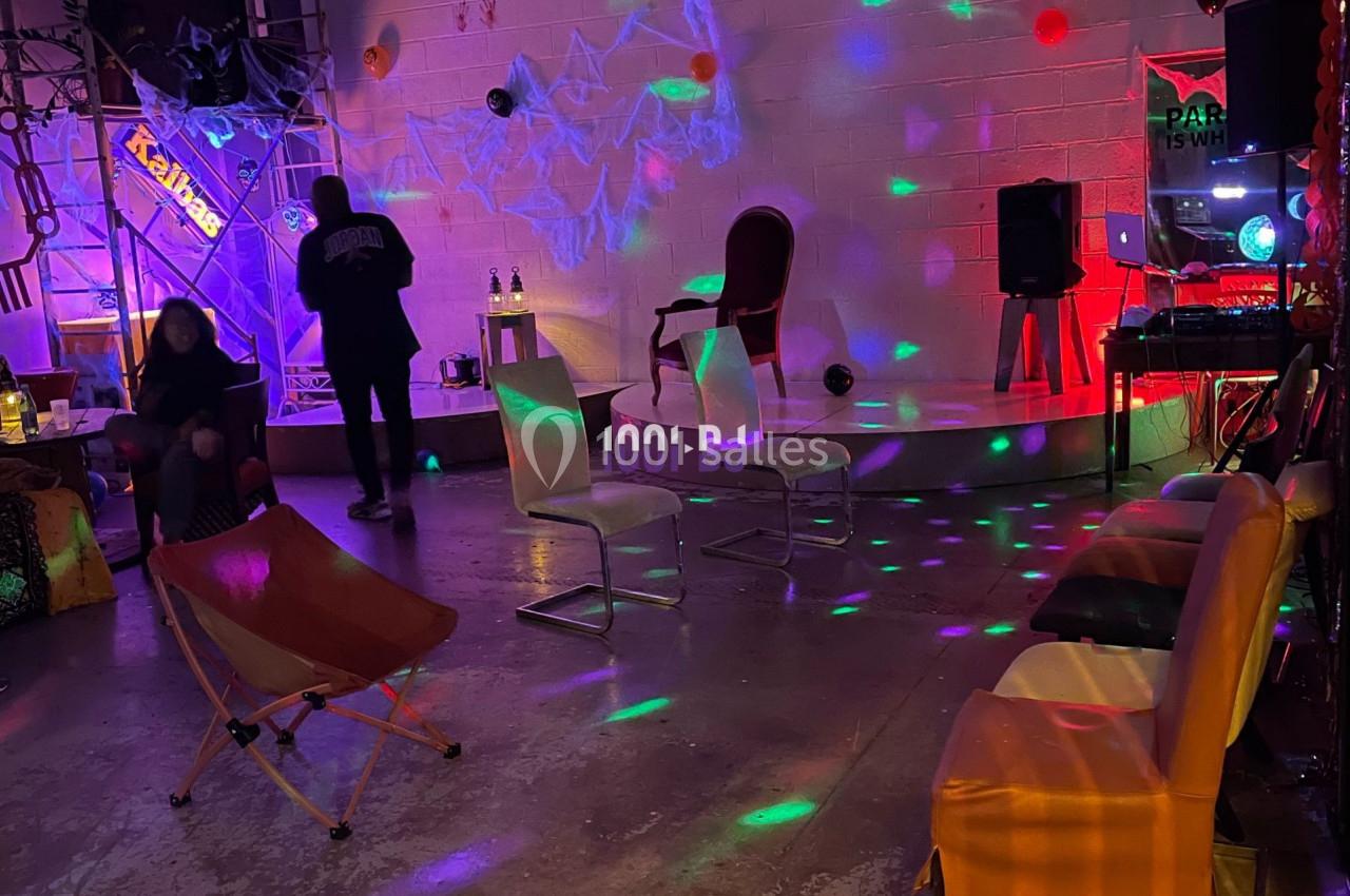 Salle industrielle avec éclairage coloré, chaises disposées, décorations et ambiance festive.