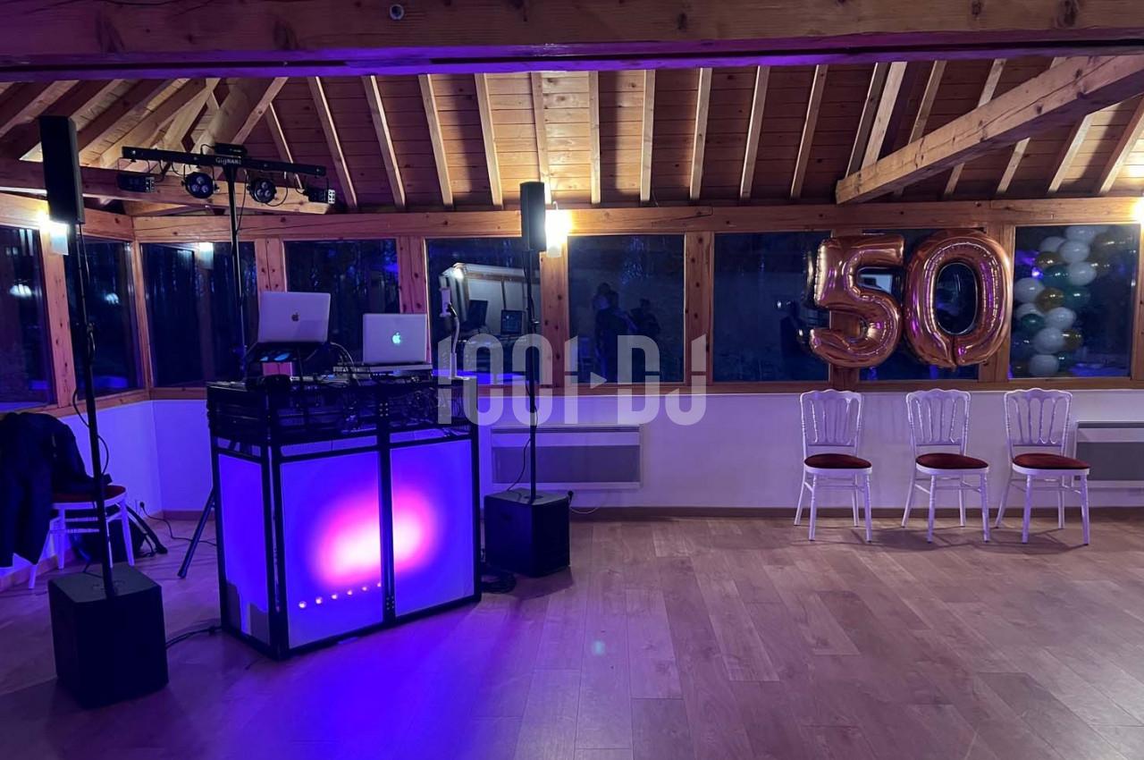 Salle décorée pour une fête, avec une table de DJ éclairée, ballons ’50’ et chaises alignées près des fenêtres.