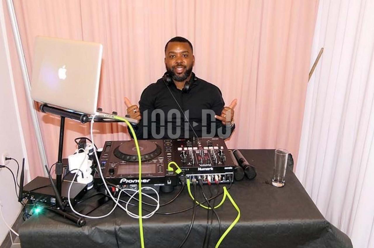 Un DJ souriant derrière une table équipée de matériel audio, avec un ordinateur portable et une table de mixage.