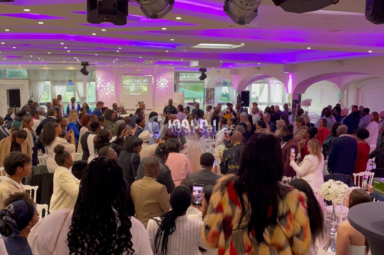 Salle de réception remplie de personnes lors d’un événement, avec éclairage violet et tables décorées.