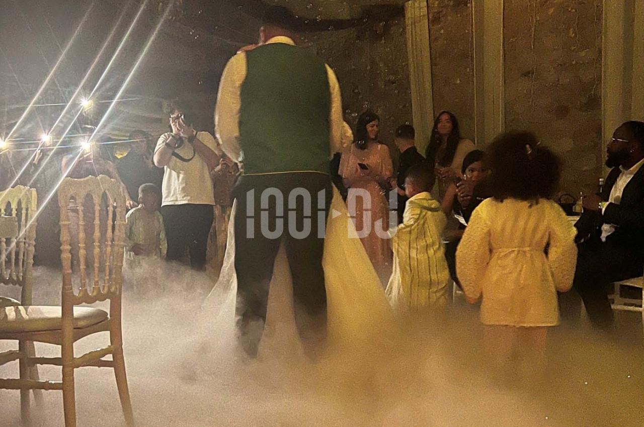 Des personnes dansent et discutent dans une salle faiblement éclairée, avec de la fumée au sol et des lumières en arrière…