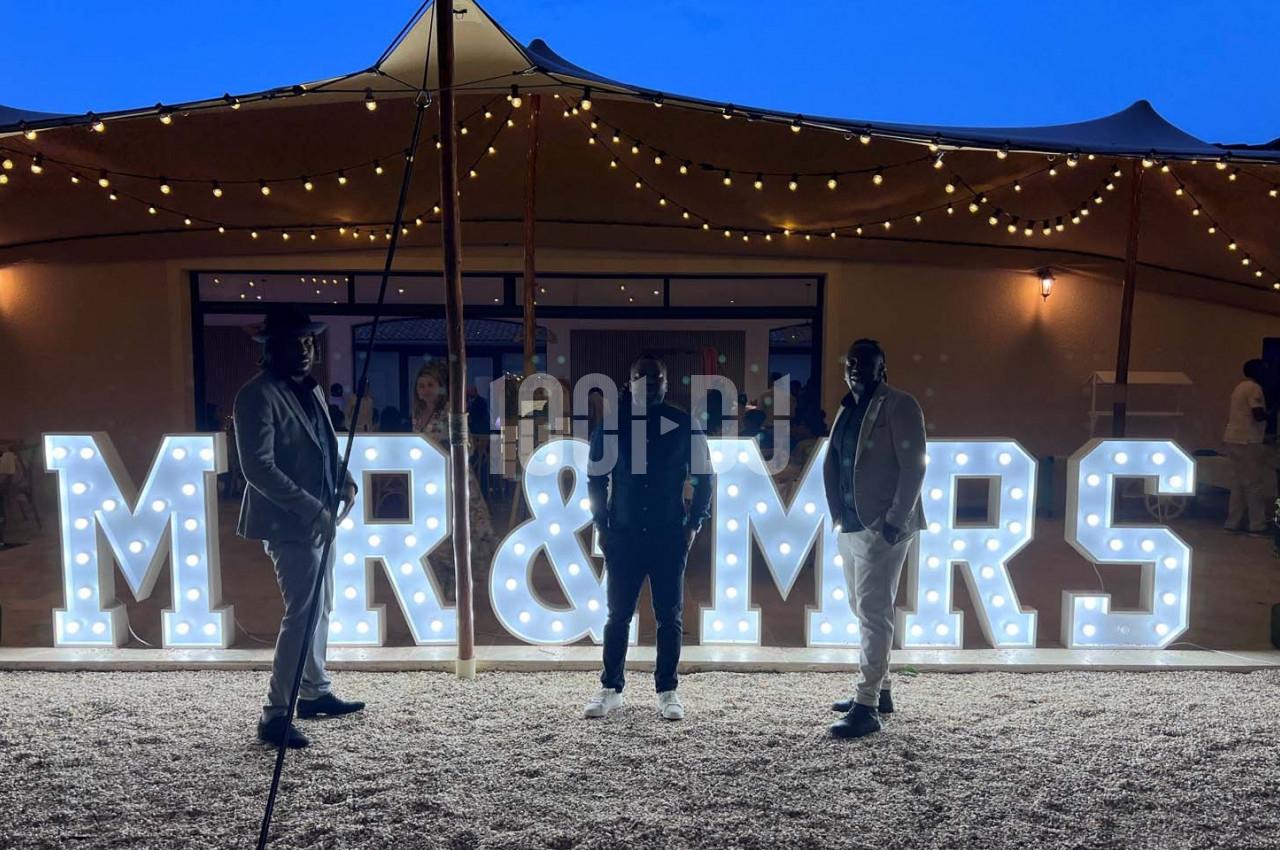 Trois personnes posent devant une enseigne lumineuse ’MR & MRS’ sous un ciel crépusculaire avec des guirlandes suspendues.