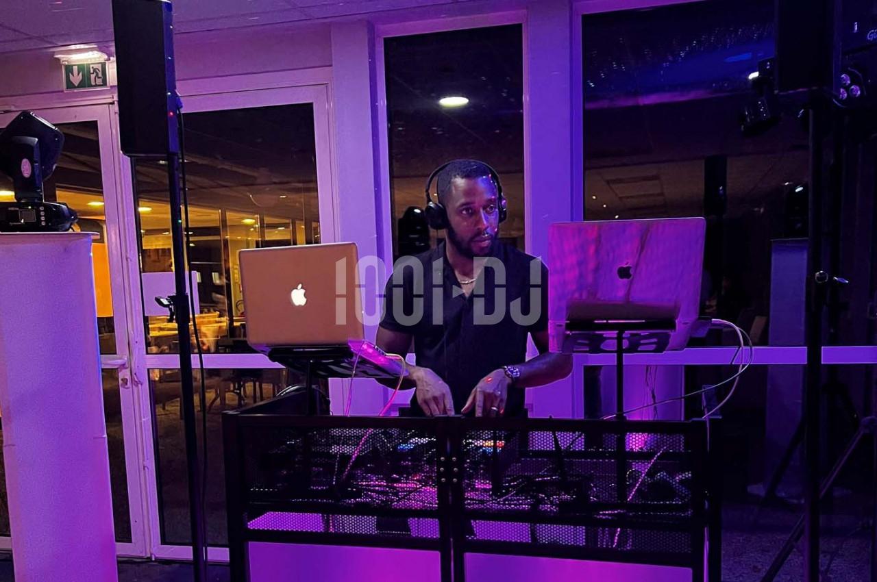 Un DJ mixe de la musique sur une table équipée d'ordinateurs et de matériel audio dans une salle éclairée en violet.