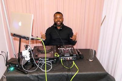 Un DJ souriant derrière une table de mixage avec un ordinateur portable et des équipements audio.