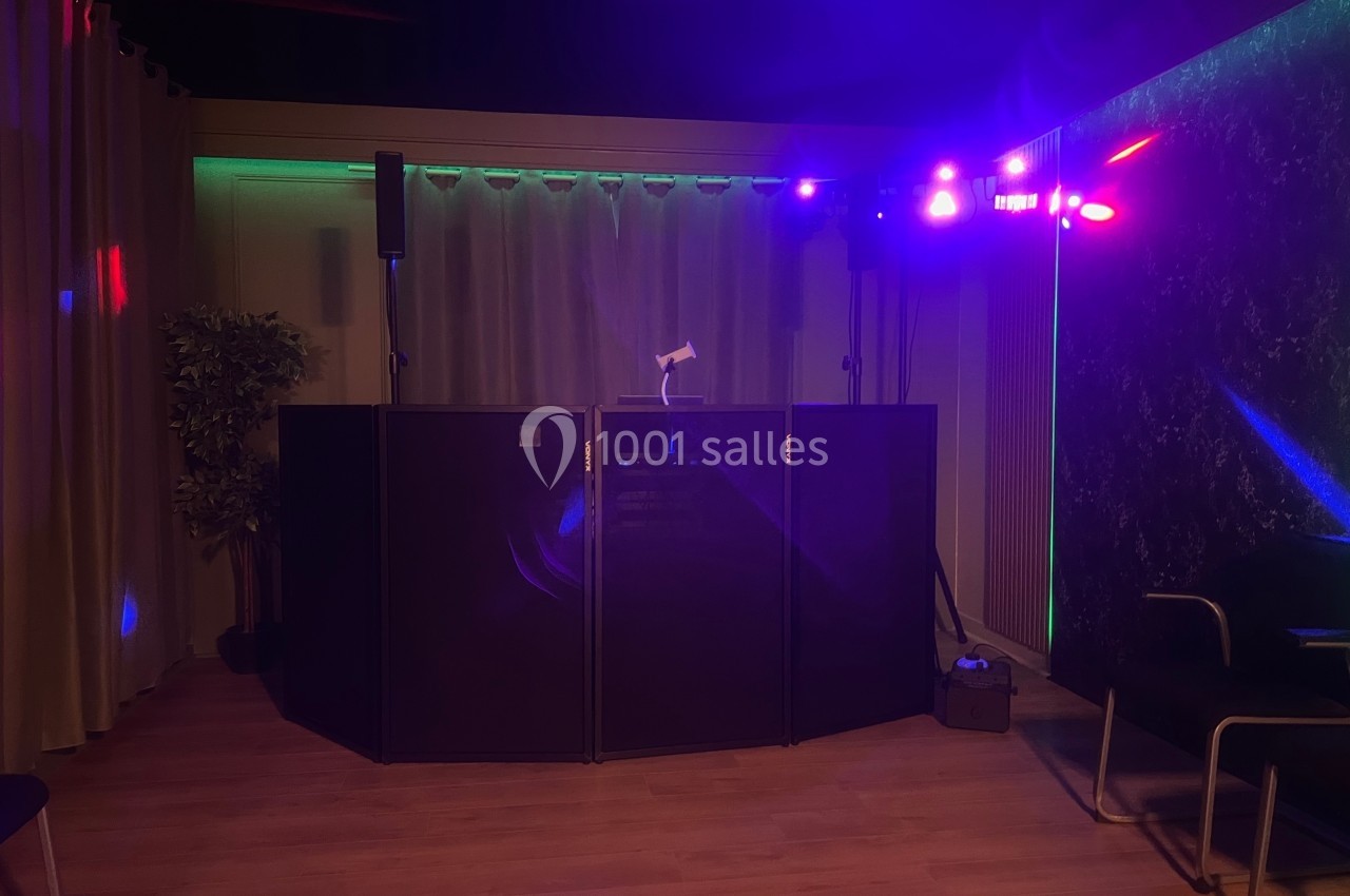 Console de DJ éclairée par des lumières colorées dans une salle sombre avec rideaux et plantes en arrière-plan.