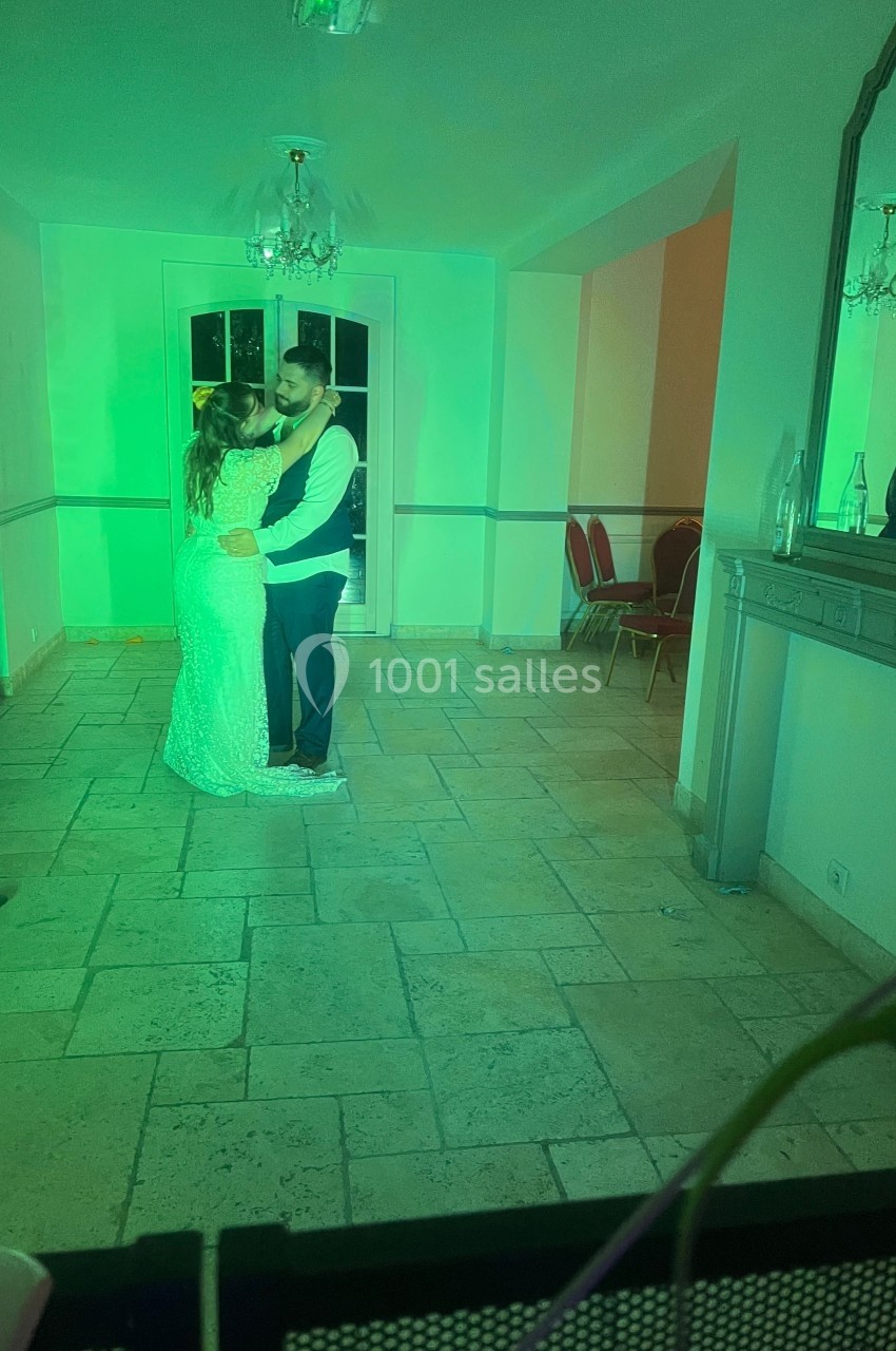 Un couple danse dans une pièce éclairée par des lumières colorées, avec un sol en pierre et des chaises alignées au fond.