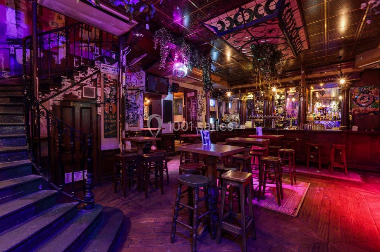 Intérieur d'un bar chaleureux avec éclairage tamisé, tables hautes, escalier en colimaçon et décoration vintage.