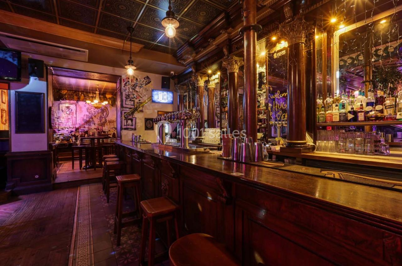 Intérieur d'un bar avec comptoir en bois, tabourets, luminaires suspendus et étagères garnies de bouteilles.