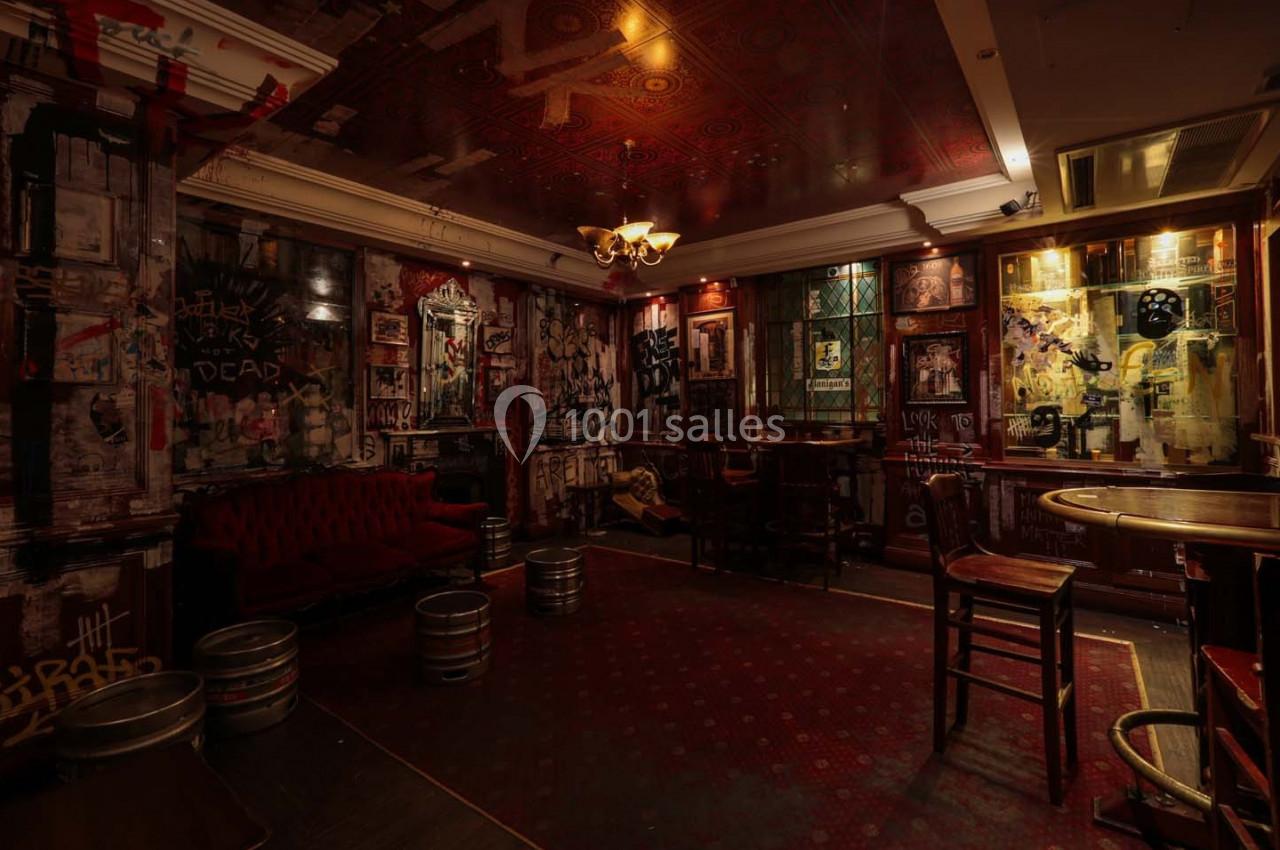 Salle sombre d'un bar avec murs couverts de graffitis, mobilier en bois, canapés rouges et tonneaux métalliques.