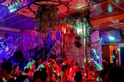 Salle animée avec des personnes dansant sous des lumières colorées, décorée de plantes suspendues et de graffitis.
