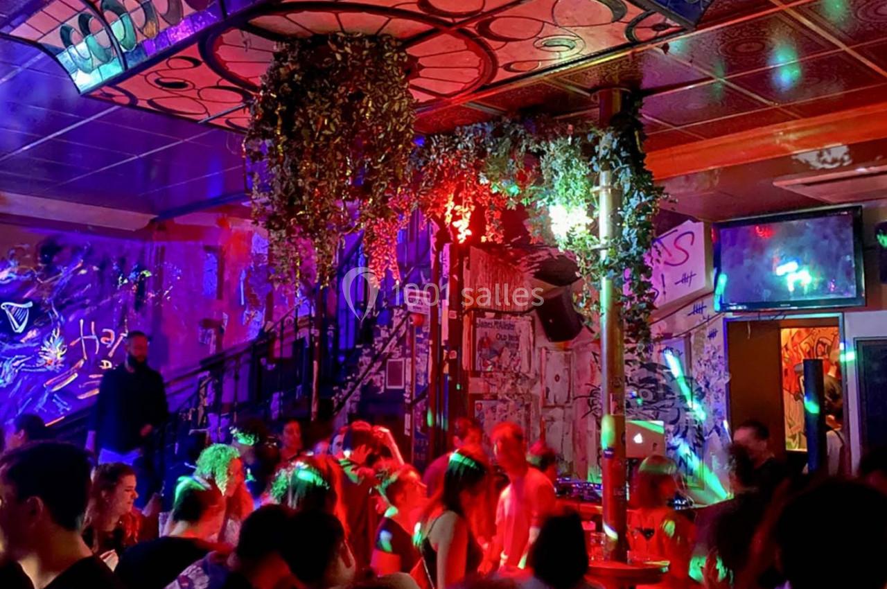 Salle animée avec des personnes dansant sous des lumières colorées, décorée de plantes suspendues et de graffitis.