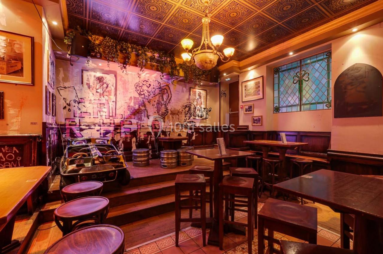 Intérieur d'un bar chaleureux avec des tables en bois, un mur décoré et une petite scène surélevée.