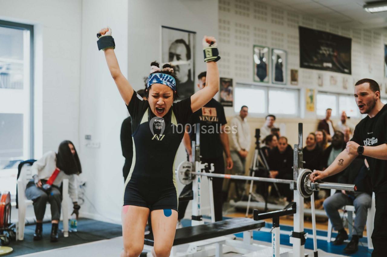Une athlète célèbre sa réussite lors d'une compétition de musculation, entourée de spectateurs dans une salle de sport.