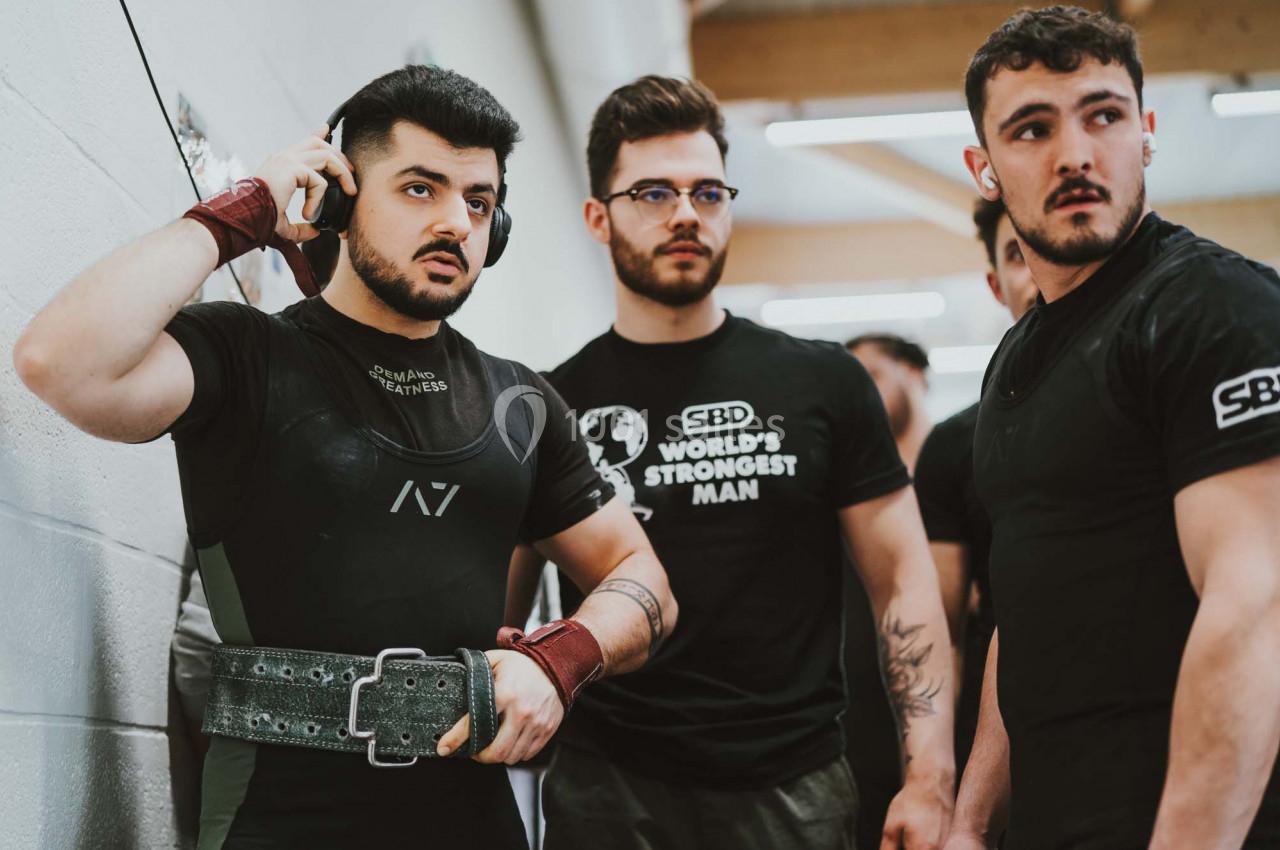 Trois hommes en tenue sportive, l'un ajustant sa ceinture de musculation, dans un environnement intérieur lumineux.