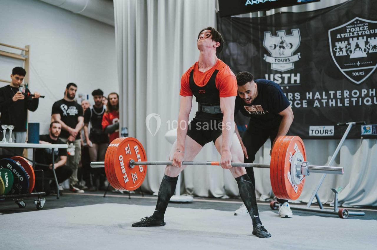 Un haltérophile soulève une barre chargée lors d'une compétition, sous le regard attentif d'un coach et du public.