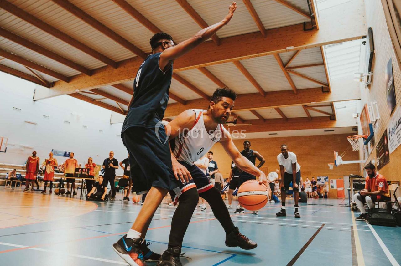 Deux joueurs de basketball s'affrontent sur un terrain intérieur, l'un dribblant tandis que l'autre défend.