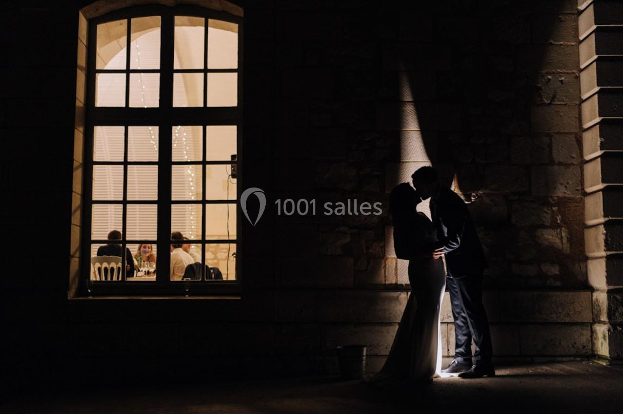 Un couple s'embrasse dans l'ombre près d'une fenêtre éclairée, révélant des convives à l'intérieur.