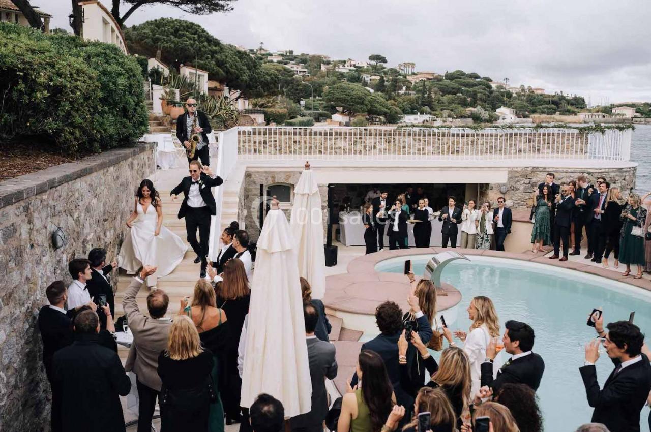 Un couple descend un escalier extérieur vers une réception près d'une piscine, entouré d'invités élégants.