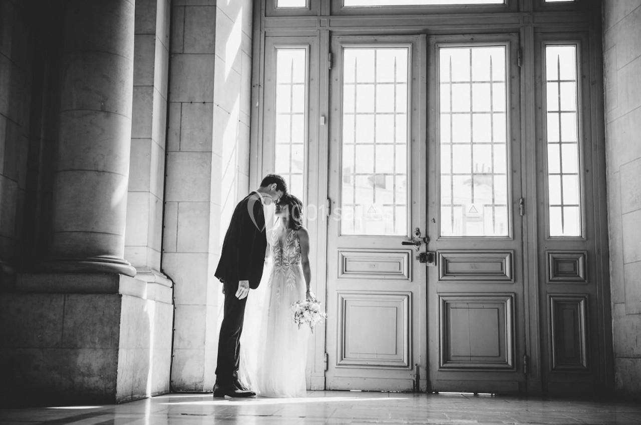 Un couple en tenue de mariage s'embrasse devant une grande porte vitrée dans un bâtiment lumineux.