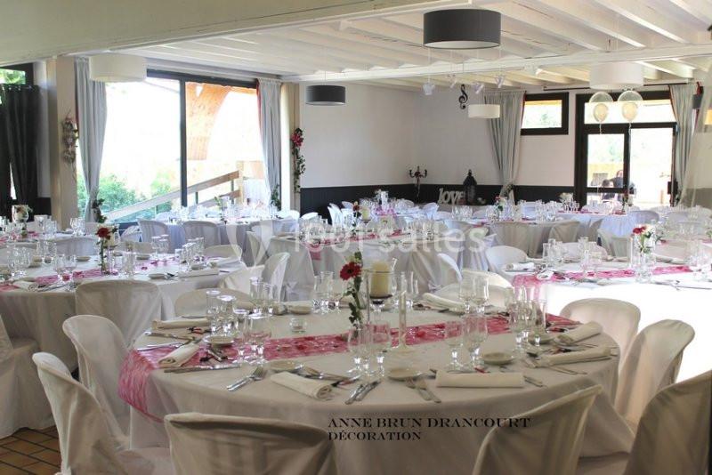 Salle de réception décorée avec des tables rondes, nappes blanches, chemins de table roses et vaisselle élégante.