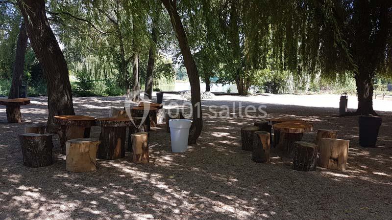 Tables et sièges en bois disposés sous des arbres dans un espace ombragé, avec vue partielle sur un plan d'eau.