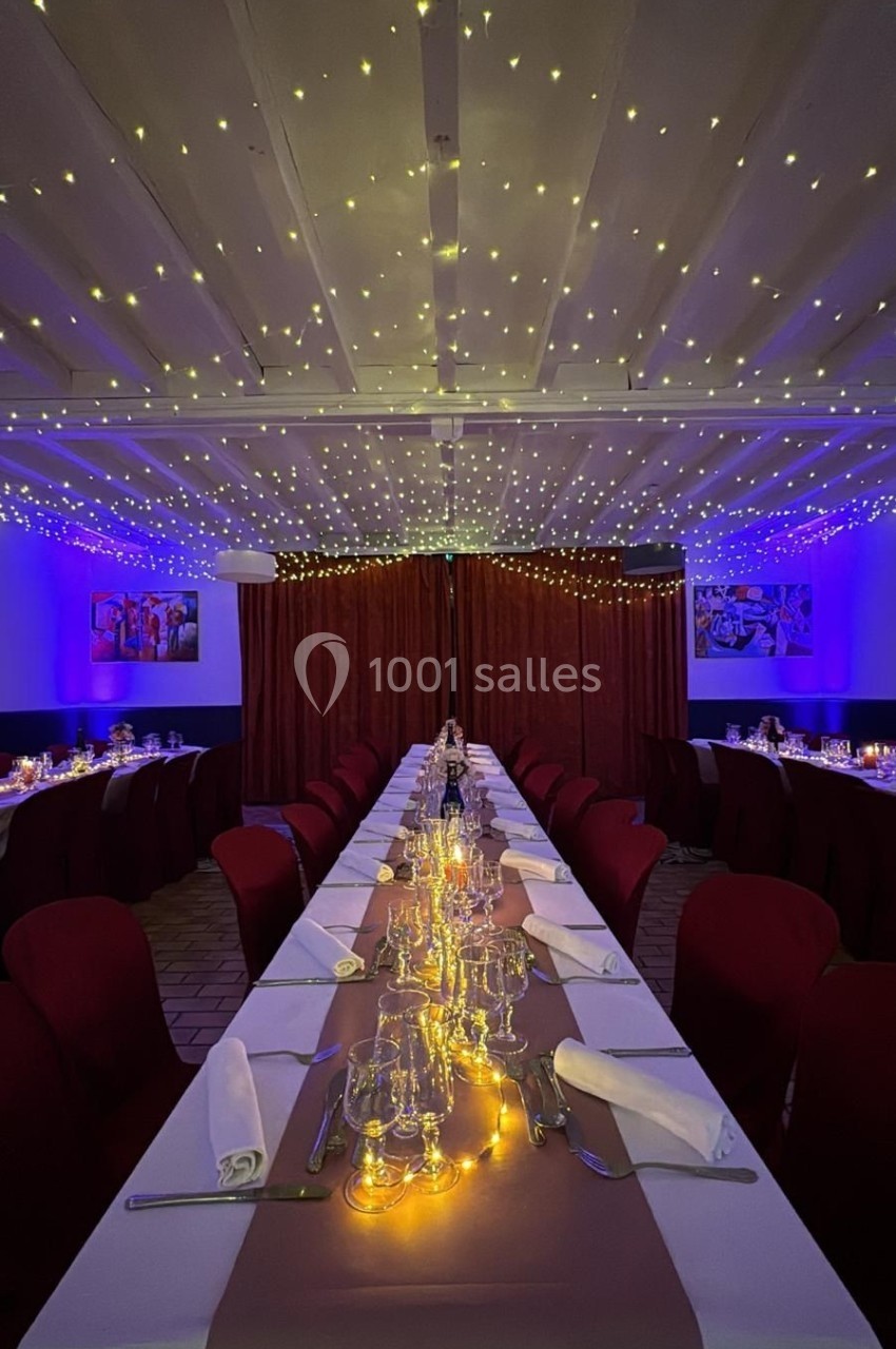 Salle de réception décorée avec une longue table dressée, nappes blanches, chaises rouges et guirlandes lumineuses au…