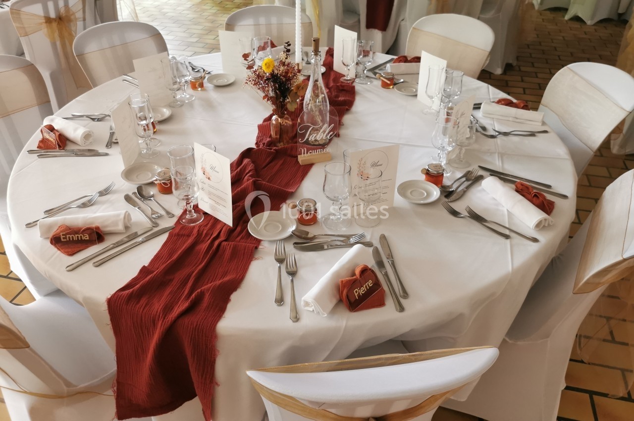 Table ronde décorée pour un événement, avec nappes blanches, chemins de table rouges, vaisselle et marque-places.