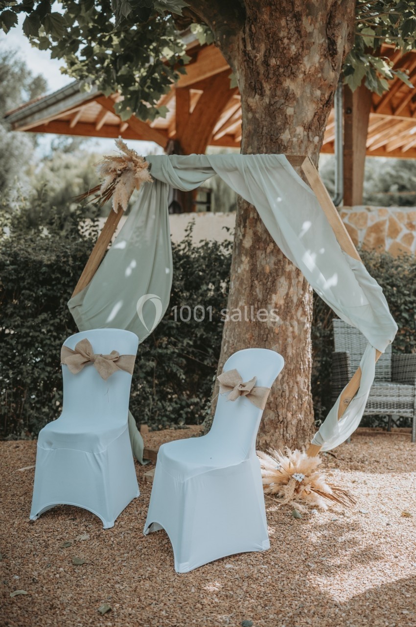 Deux chaises blanches décorées de nœuds en toile de jute devant un arbre orné de tissu vert clair et de fleurs séchées.
