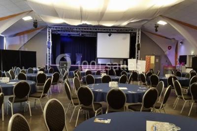 Salle de réception aménagée avec des tables dressées pour un événement, éclairage tamisé et scène en arrière-plan.