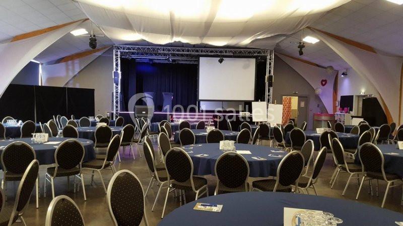 Salle de réception avec tables rondes dressées, scène équipée d'un écran et d'un éclairage.