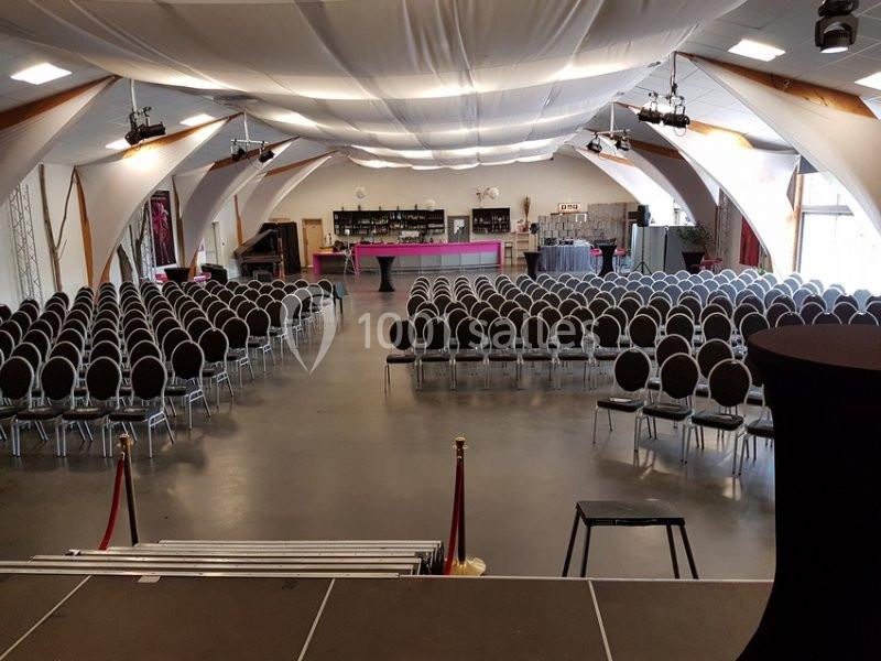 Salle de réception avec rangées de chaises alignées face à une scène, bar en arrière-plan.