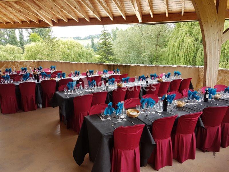 Salle en extérieur avec tables dressées, nappes noires, chaises rouges et serviettes bleues, entourée de verdure.
