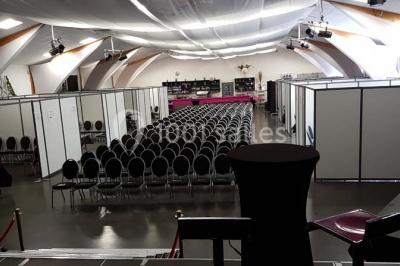 Salle de réception aménagée avec des tables dressées pour un événement, éclairage tamisé et scène en arrière-plan.