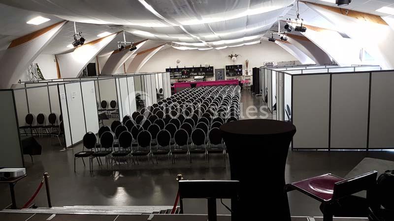 Salle aménagée avec des rangées de chaises noires et des cloisons modulaires, éclairée par des spots au plafond.