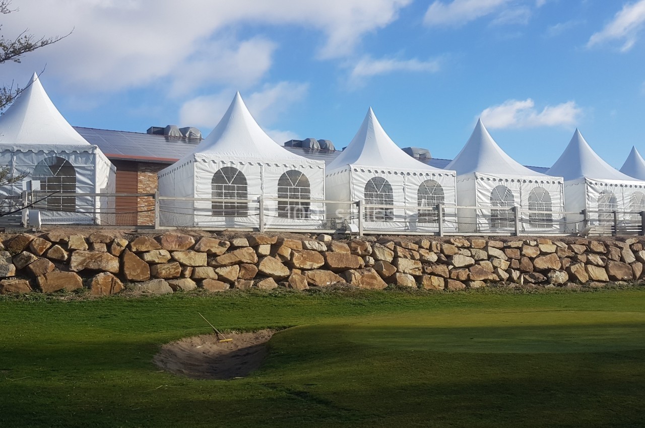 Tentes blanches alignées sur une terrasse surélevée en pierre, entourées de pelouse et sous un ciel partiellement nuageux.
