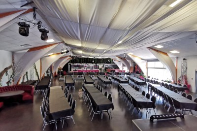 Salle de réception aménagée avec des tables dressées pour un événement, éclairage tamisé et scène en arrière-plan.