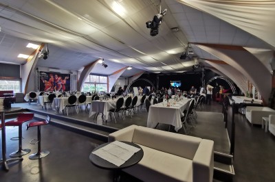 Salle de réception aménagée avec des tables dressées pour un événement, éclairage tamisé et scène en arrière-plan.