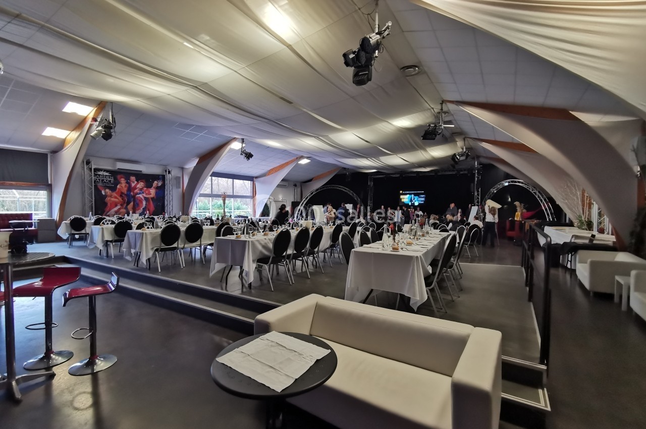 Salle de réception aménagée avec des tables dressées pour un événement, éclairage tamisé et scène en arrière-plan.