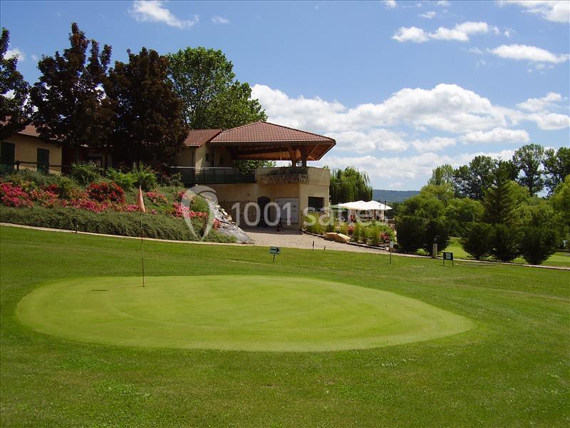 Vue d'un terrain de golf avec un green au premier plan et un bâtiment entouré de végétation en arrière-plan.