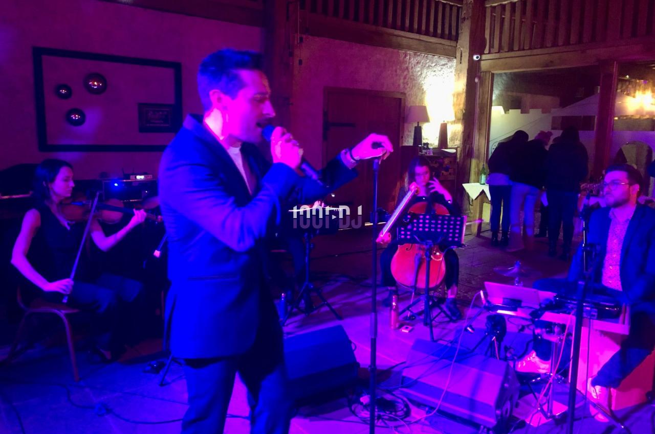 Un chanteur en costume interprète sur scène, accompagné de musiciens jouant du violon, violoncelle et clavier.